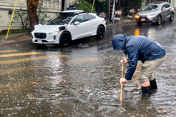 A Mission District resident attempts to unclog a few storm drains on a flooded street as a driverless Waymo taxi passes in the background, Friday, Nov. 22, 2024, in San Francisco. (AP Photo/Haven Daley)