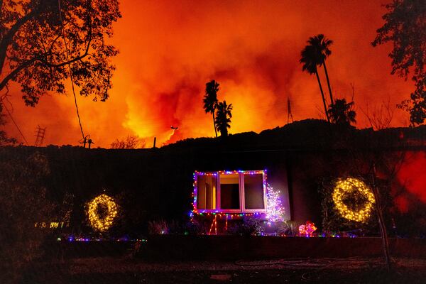 A helicopter drops water on the Palisades Fire behind a home with Christmas lights in Mandeville Canyon, Friday, Jan. 10, 2025, in Los Angeles. (AP Photo/Ethan Swope)