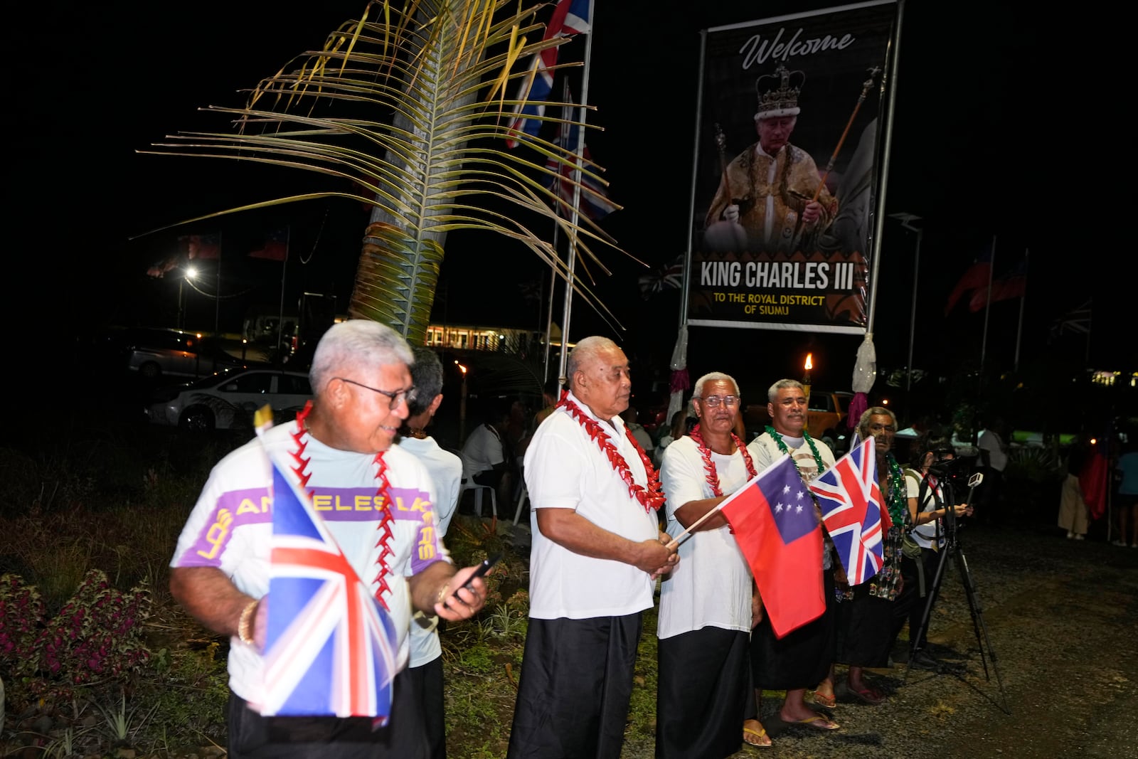 Men hold flags as they wait for the arrival of Britain's King Charles III and Queen Camilla in the village of Siumu, Samoa, on Wednesday, Oct. 23, 2024. (AP Photo/Rick Rycroft)