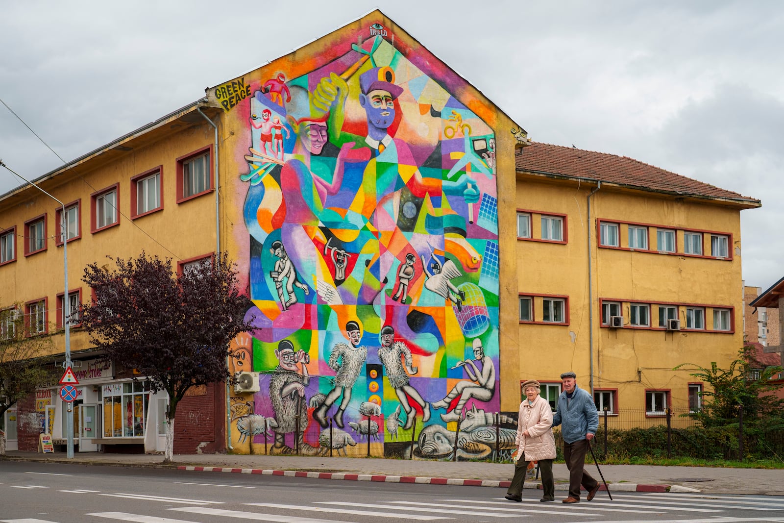 People cross a street backdropped by a mural by Romanian artist IRLO in Petrosani, southern Romania, Friday, Oct. 11, 2024. The mural depicts the evolution of the Jiu Valley, Romania's coal mining region as steps are taken to move away from coal based energy production to green energy solutions. (AP Photo/Vadim Ghirda)