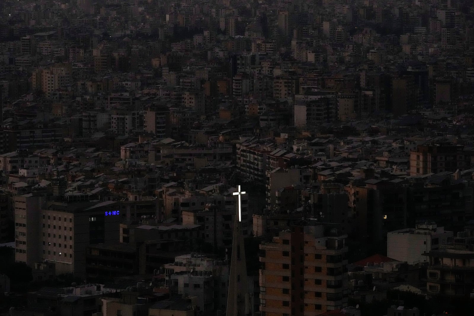 The cross atop of Our Lady of Hadath Church appears in front Dahiyeh suburb, background, that remains in darkness after Israeli airstrikes, Lebanon, Wednesday, Oct. 16, 2024. (AP Photo/Hassan Ammar)