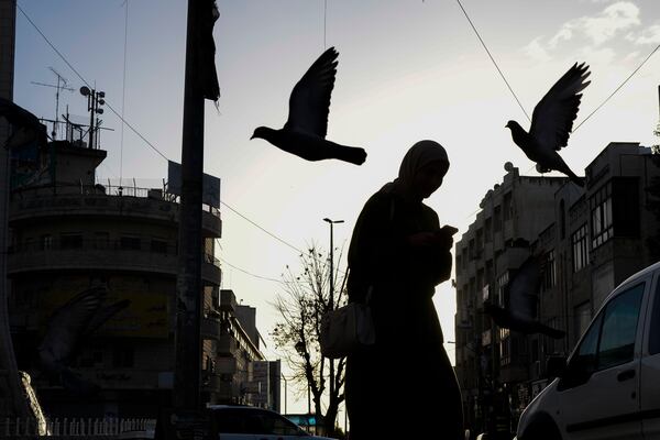 A woman walks amid pigeons flying in a square in the West Bank city of Ramallah, Friday, Dec. 27, 2024. (AP Photo/Matias Delacroix)