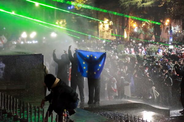 Demonstrators with an EU flag stand atop of a tomb under running water from a water cannon rallying outside the parliament's building to continue protests against the government's decision to suspend negotiations on joining the European Union in Tbilisi, Georgia, on Monday, Dec. 2, 2024. (AP Photo/Zurab Tsertsvadze)